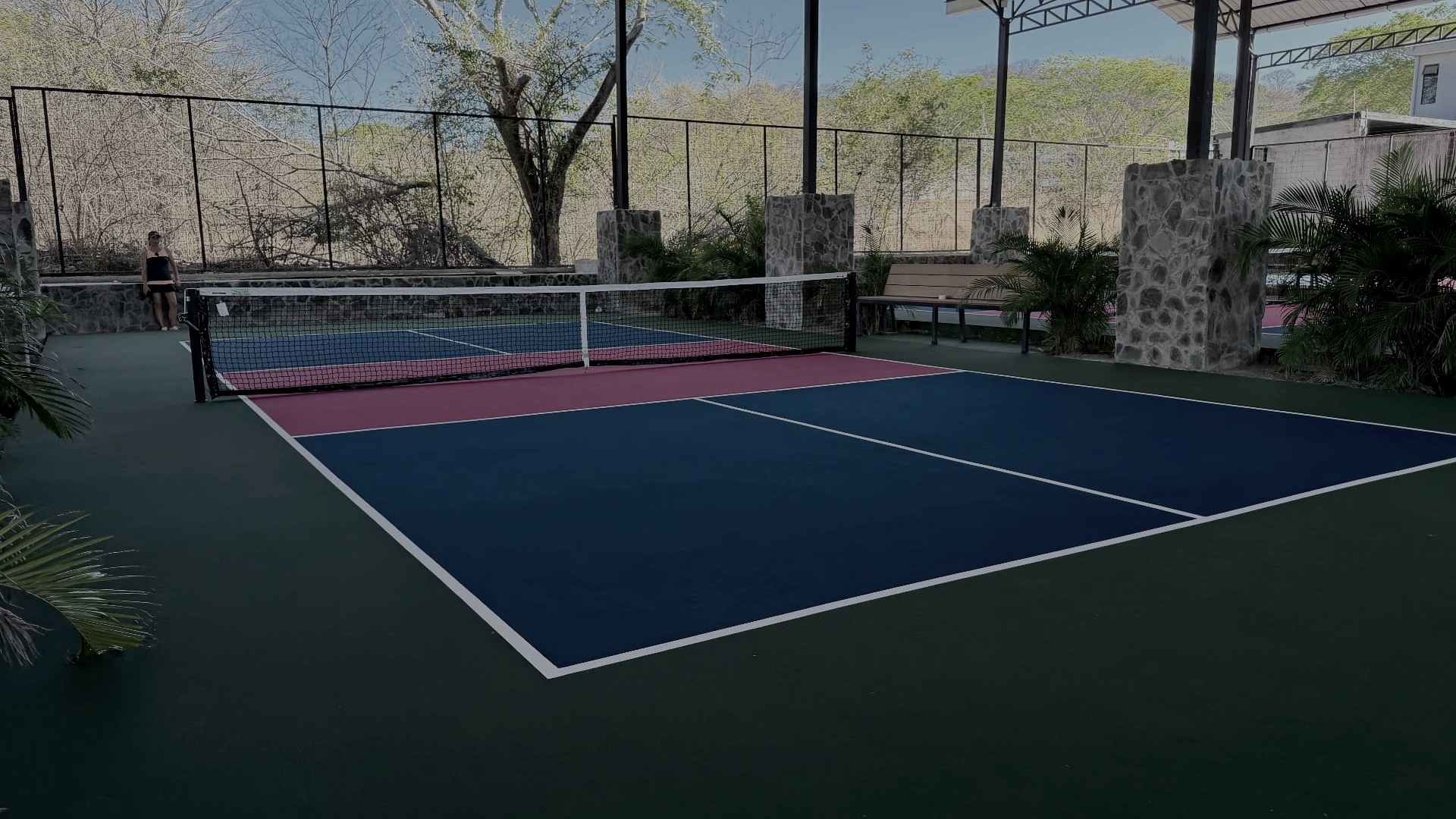Covered pickleball courts at Finca Escondida, blue playing surfaces with magenta non-volley zones under an arched roof open to tropical forest on both sides